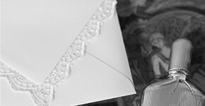 SAY WOW Karina von Ende Detailaufnahme einer weißen Hochzeitseinladung mit Spitzenrand neben einem Parfümflakon und Ringen, Schwarz-Weiß